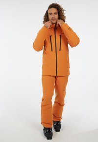 Chaqueta de esquí impermeable naranja con cremalleras negras, dos bolsillos en el pecho y pantalones a juego. Tejido texturizado, con capucha y un ajuste elegante.