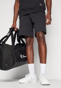 Shorts de sport noirs au design structuré, associés à des baskets blanches et à un sac de sport noir arborant le logo Champion.