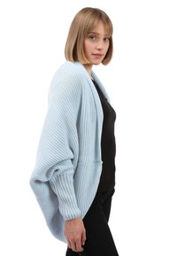 Hellblauer, übergroßer Strickcardigan mit gerippter Textur, weiten Ärmeln und vorderen Taschen, getragen über einem schwarzen Oberteil.
