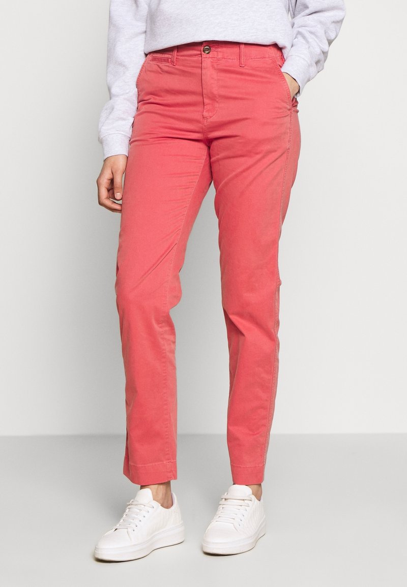 Personne portant un pantalon corail à jambe droite, des baskets blanches et un sweat-shirt gris clair, debout avec une main dans la poche.