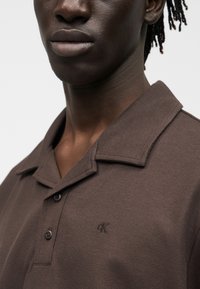 Κοντινό πλάνο ενός ατόμου που φορά ένα καφέ πόλο με υφή, γιακά και μικρό λογότυπο «ck» στο στήθος.