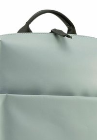 Sac à dos léger et doux de couleur gris clair fabriqué en tissu texturé, doté d'une petite poignée en haut et d'une poche avant saillante avec une fermeture éclair.