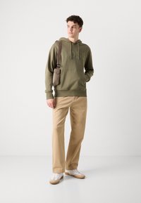 Felpa verde oliva con logo in rilievo, pantaloni beige e sneakers bianche con dettagli color tabacco, indossati da una persona con una borsa a spalla marrone.
