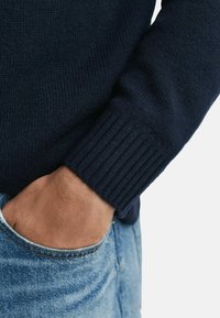 Námornícky pletený sveter s žebrovanými manžetami, spárovaný s svetlomodrými džínsami. Má priliehavý strih s textúrovanou tkaninou a neformálny vzhľad.