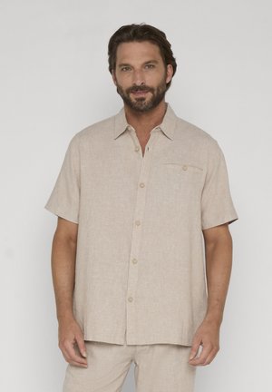 Mann mit Bart trägt beigefarbenes kurzärmeliges Hemd mit Knöpfen und passende Hose, steht vor schlichtem hellem Hintergrund.