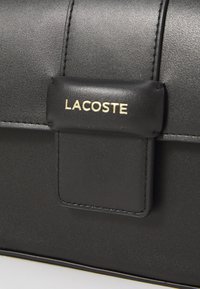 Svart skinnveske med en glatt tekstur, med en gull «Lacoste»-logo og klaffelukking. Synlig søm fremhever kantene.