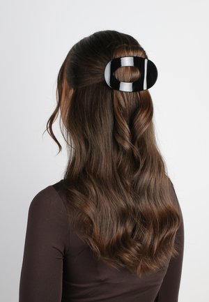 Fermaglio nero lucido con un design ovale, che tiene fermo i capelli castani mossi, dotato di un foro circolare centrale. Superficie liscia e riflettente.