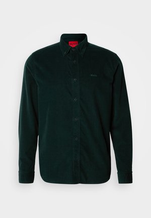 Tumši zaļš korduroja krekls ar garām piedurknēm, ar pogām aizdaram un melnām pogām. Uz krūtīm ir smalks zīmola logotips un sarkans birka.