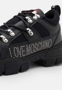 Svart sneaker med läder- och meshmaterial, silverfärgad dekorativ hårdvara och nitar med texten "LOVE MOSCHINO" på sidan. Texturerad sula.