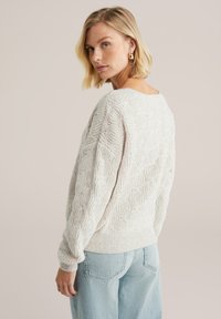 Maglione lavorato a maglia grossa di colore crema chiaro con motivo a treccia testurizzato, maniche raglan e polsini a coste. Abbinato a jeans a vita alta di colore blu chiaro.