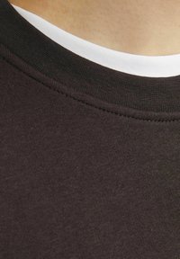 Donkerbruine T-shirt met een contrasterende zwarte geribbelde kraag, met daaronder een gelaagde witte T-shirt met ronde hals zichtbaar. Zachte katoen textuur.