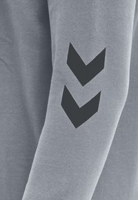 Graues Langarmshirt mit glatter Textur, das ein großes schwarzes Chevron-Motiv am linken Ärmel aufweist. Einfaches Design, keine Muster.