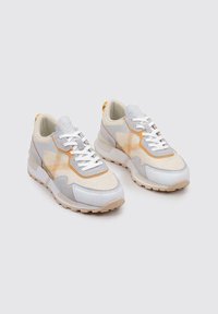 Las zapatillas presentan una parte superior de malla en beige y gris con acentos naranjas, cordones blancos y una suela gruesa de color crema. Los paneles texturizados añaden detalle.