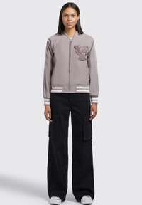 Veste bomber grise claire avec un logo de tigre sur la poitrine, des poignets et un ourlet rayés, portée avec un pantalon cargo noir et des baskets blanches.