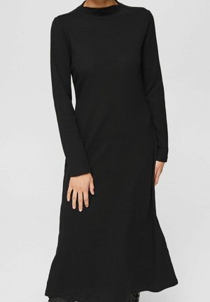 Kobieta ubrana w czarną, prążkowaną sukienkę midi z długim rękawem i wysokim kołnierzem o prostej sylwetce na jednolitym szarym tle.