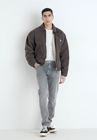 Chaqueta bomber marrón, camiseta blanca, vaqueros grises, zapatillas negras. La chaqueta tiene una cremallera y bolsillos laterales, los vaqueros tienen una textura desgastada.