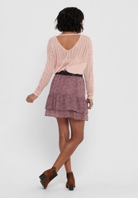 Pull en tricot rose clair avec un dos ouvert, jupe florale superposée dans des nuances de rose, taille noire, et bottines marron à talon carré.