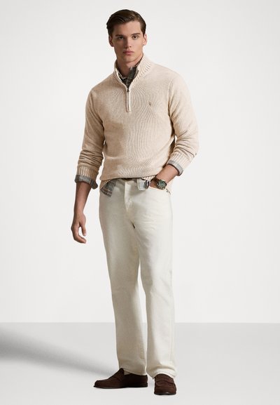 Beige gebreide trui met een halve ritskraag, gedragen over een geruit overhemd. Gecombineerd met ivoorkleurige broek en bruine loafers. Gladde textuur.