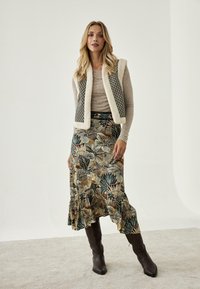 Top de punto beige debajo de un chaleco de piel sintética con un detalle de estampado, combinado con una falda maxi de corte acampanado con estampado floral y botas marrones hasta la rodilla.