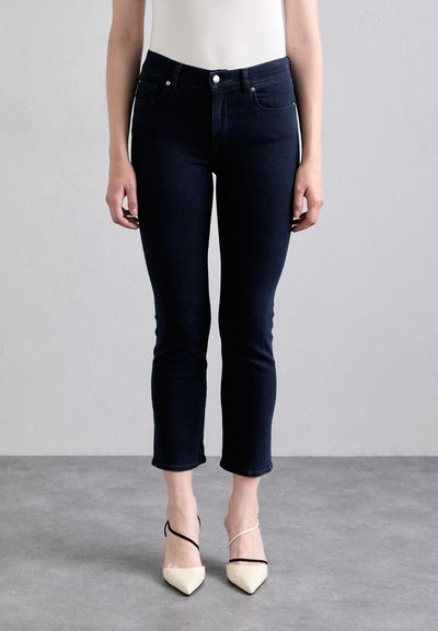 Jeans închis la culoare, slim fit, cu talie înaltă, lungime până la glezne și design cu cinci buzunare, asortați cu pantofi cu toc ascuțit de culoare bej cu curele negre.