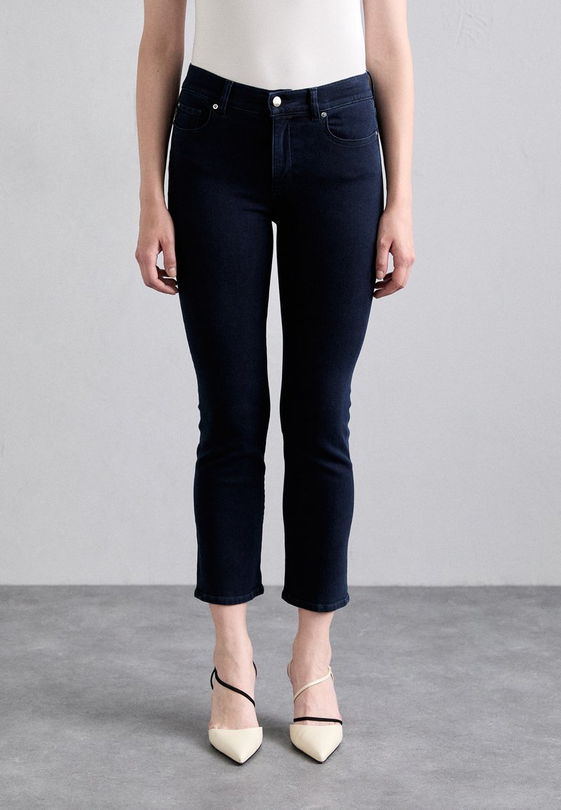 Jeans ajustés bleu marine à taille haute, longueur cheville, et design à cinq poches, associés à des talons beiges à bouts pointus avec des brides noires.