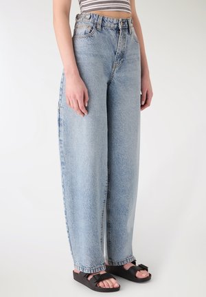 Persona che indossa jeans a vita alta, di colore azzurro chiaro, con gambe larghe e sandali neri con fibbia, in piedi su uno sfondo bianco uniforme.