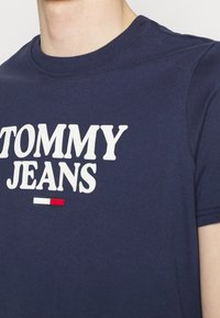 Tmavě modré bavlněné tričko s bílým logem "TOMMY JEANS" a malým červeným a bílým akcentovým obdélníkem pod textem. Klasický výstřih ve stylu crew.