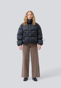 Žena s kudrnatými vlasy oblečená v černé péřové bundě, širokých hnědých kalhotách a černých botách, stojící na světle šedém pozadí.