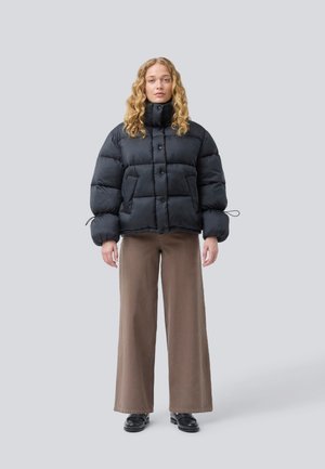 Zwarte gewatteerde jas met een hoge kraag, met prominente knoppen, elastische manchetten en een boxy silhouette. Gecombineerd met bruine wijd uitlopende broeken.