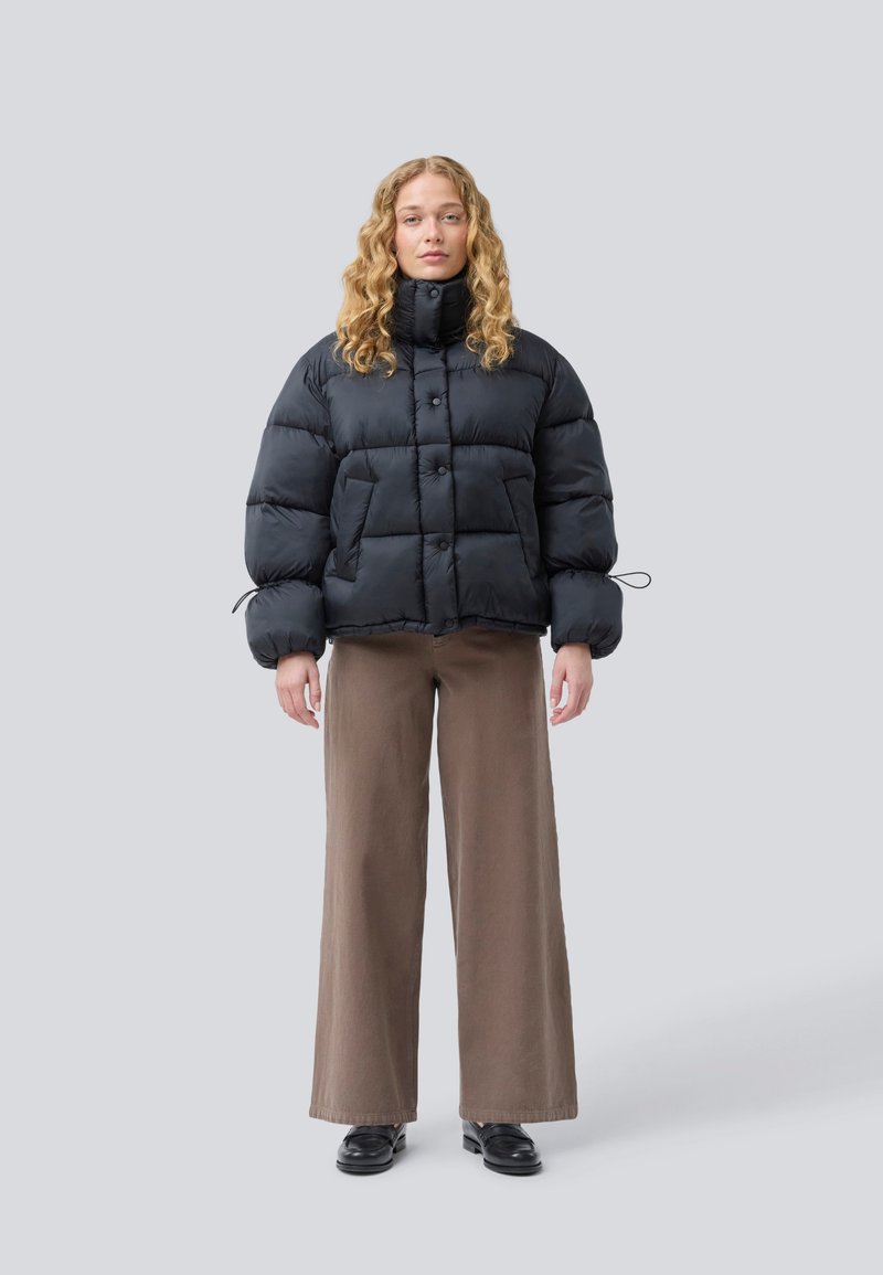 Žena s kudrnatými vlasy oblečená v černé péřové bundě, širokých hnědých kalhotách a černých botách, stojící na světle šedém pozadí.