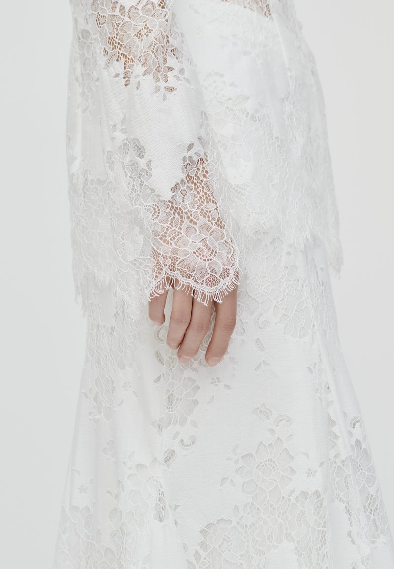 Weißes Spitzenkleid mit floralen Mustern, langen Ärmeln mit kunstvollen Spitzenabschlüssen und einem glatten Stoffuntergrund. Hand sanft an der Seite abgelegt.