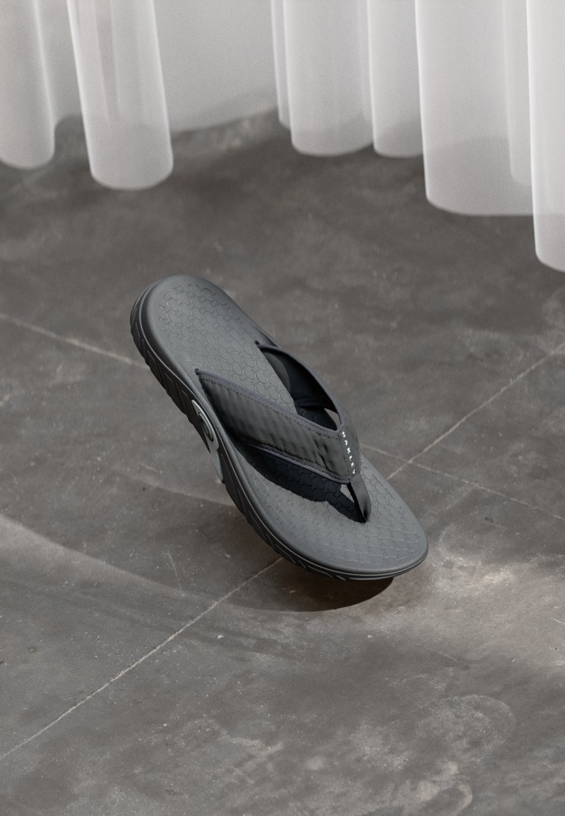 Ein einzelner schwarzer Flip-Flop-Sandal mit strukturiertem Sohle, der auf einem grauen Fliesenboden unter durchscheinenden weißen Vorhängen liegt.