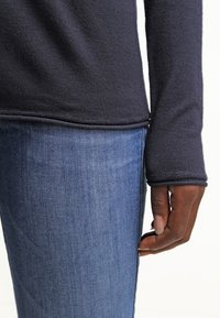 Chemise à manches longues bleu marine foncé avec un tissu texturé, associée à un jean bleu ajusté. La chemise présente un ourlet droit et des manches ajustées.