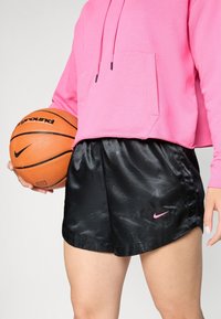 Pantalones cortos de baloncesto de satén negro con un patrón sutil, combinados con una sudadera corta rosa y sosteniendo un balón de baloncesto naranja.