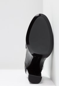 Semelle de chaussure habillée noire brillante avec un talon carré, inclinée contre un fond blanc d'angle.