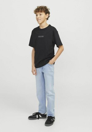 T-shirt nera a maniche corte con un accento rettangolare nero sul petto, abbinata a jeans azzurri chiari e sneakers nere.