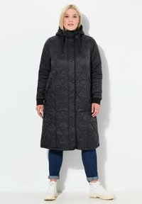 Svart vadderad lång jacka med huva, med ett diamantmönster, framknappar och sidofickor, kombinerad med jeans och vita sneakers.