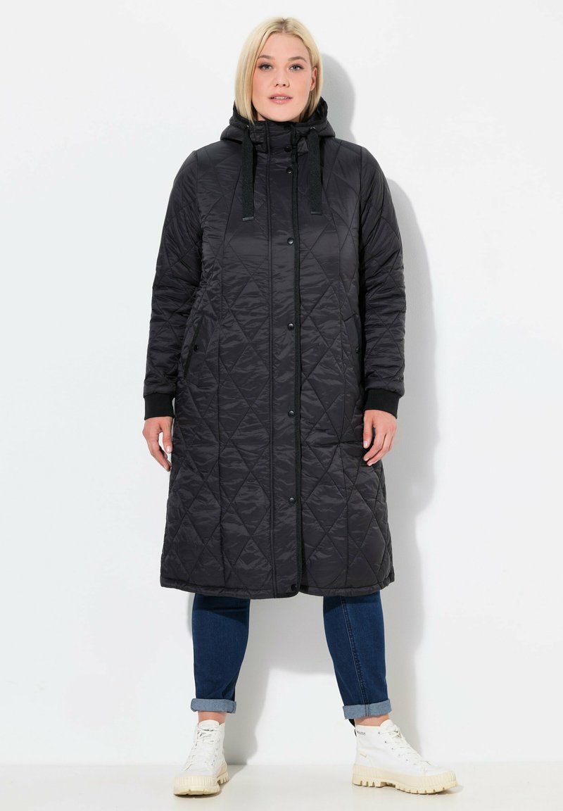 Svart vadderad lång jacka med huva, med ett diamantmönster, framknappar och sidofickor, kombinerad med jeans och vita sneakers.