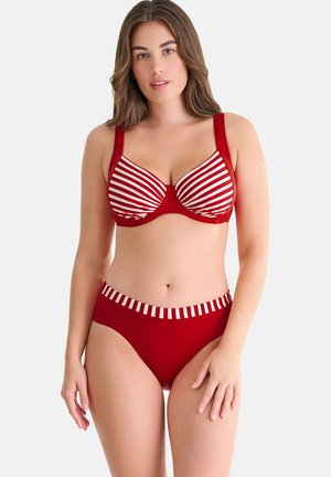 Vrouw die een rood-wit gestreepte beha draagt met bijpassende rode slip met een tailleband met witte strepen, staand tegen een effen achtergrond.