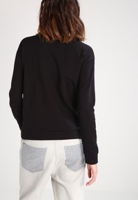 Sudadera negra de manga larga con cuello redondo y dobladillo acanalado, combinada con jeans claros que destacan por sus bolsillos en gris.