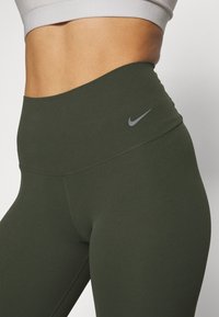Leggings taille haute vert olive avec une texture lisse et extensible. Dotés d'un logo Nike gris sur la ceinture. Détail de couture soigné.