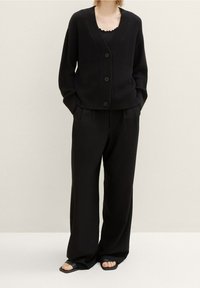 Musta ribbineuletakki V-aukkolla ja etupuolen napituksella, yhdistetty korkeavyötäröisiin mustiin housuihin. Minimalistinen muotoilu, rento istuvuus.