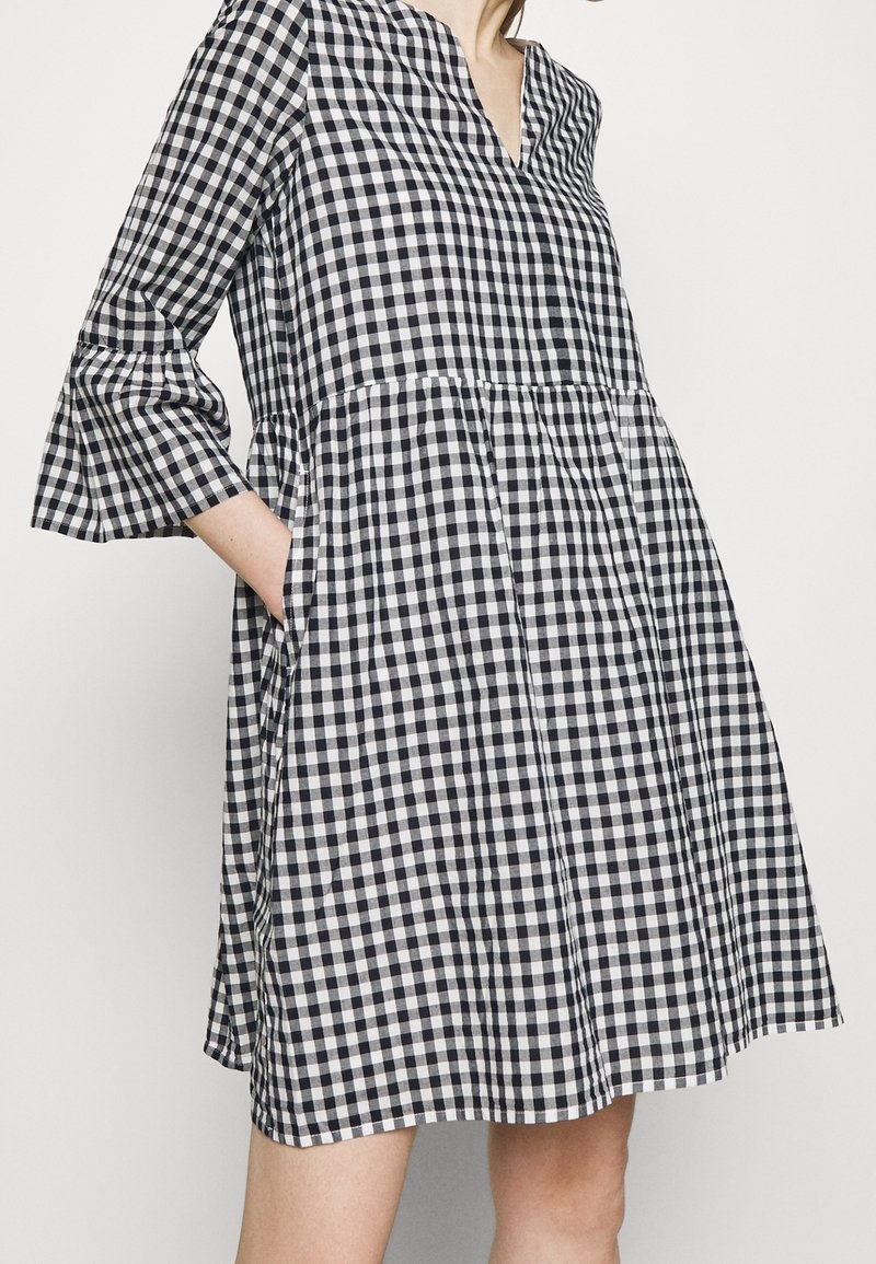 Femme portant une robe à carreaux vichy noir et blanc avec des manches cloche 3/4 et des poches latérales, debout devant un fond clair uni.