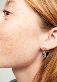 Pendientes de plata con tachuelas y un acento de perla, con una gema cuadrada oscura colgando debajo, sobre un cutis con pecas.