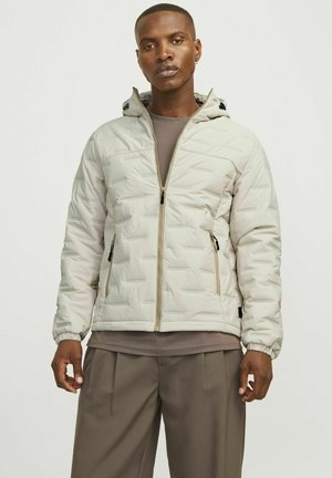 Veste matelassée beige clair avec fermeture éclair à l'avant, texture à motifs, poignets élastiques et deux poches zippées sur les côtés. Design avec capuche et doublure grise.