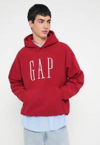 Felpa rossa con cappuccio e logo "GAP" bianco, realizzata in tessuto morbido. Dotata di tasca frontale e sovrapposta a una camicia a righe blu sottostante.