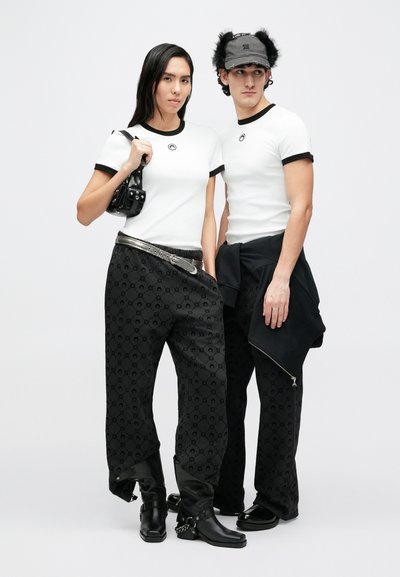 Dva modelové mají na sobě sladěné outfity: bílé trička s černým lemováním, volné černé kalhoty s vzorem a černé doplňky s kovovými akcenty.