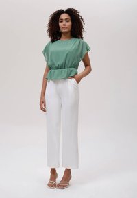 Mulher com cabelo cacheado, vestindo uma blusa verde de mangas curtas, calças brancas de perna larga e sandálias de salto brancas, de pé contra um fundo simples.