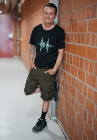 Camiseta negra con un gráfico de onda sonora, pantalones cortos verde oliva y zapatillas negras. Un hombre se apoya contra una pared de ladrillos rojos en un pasillo.