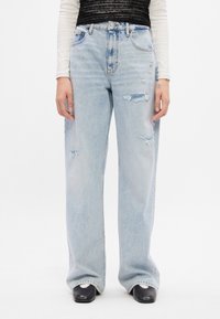 Jeans de tiro alto, de pierna ancha, en azul claro con detalles desgastados. Fabricados de mezclilla, con un leve desvanecimiento. Llevados con zuecos negros.